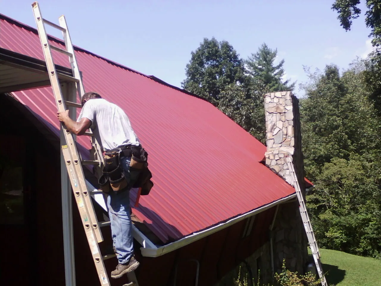 Roofer on a ladder beside a red metal roof and stone chimney — Soffit & Fascia Repair by Roofing Brothers in North Bergen